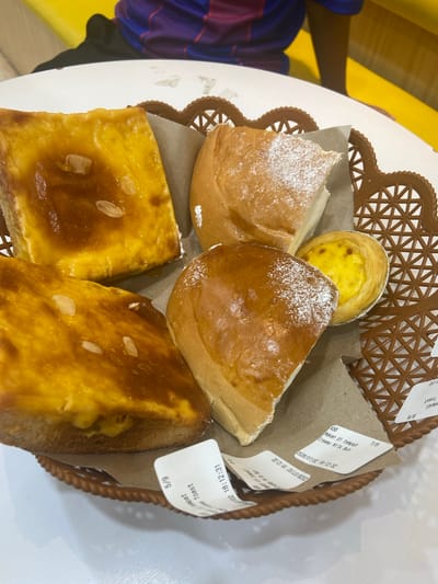 In the city of Kembang Harum, Indonesia, a brown woven basket sits on a white table, filled with baked goods. The basket contains two rectangular slices of toast topped with a golden, glistening glaze and what appear to be slivered almonds. Beside the toast are two portions of bread rolls, one of which is dusted with powdered sugar. A single egg tart, with its flaky pastry and bright yellow custard filling, is nestled amongst the rolls. Several small paper receipts are scattered around the items in the basket, one of which displays "Makan di tempat" (Eat in) and "Creamy Milk Bun" along with a date and time. In the background, a person wearing a purple and blue striped shirt is visible, and a bright yellow surface is also present.