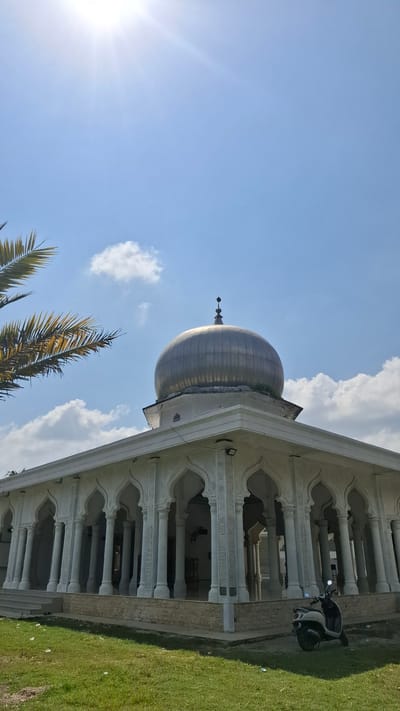 In Grong-grong, Indonesia, a bright, sunny day illuminates a mosque with white walls and a prominent silver dome. The mosque features an arcade of white, arched colonnades supporting the roof. In the foreground, a lush green lawn is visible, with a few scattered pieces of white debris. A white scooter is parked on the grass to the right of the mosque. Palm fronds from a tree frame the upper left corner of the image against a clear blue sky dotted with white, fluffy clouds. The sun is high and bright, casting a strong light.