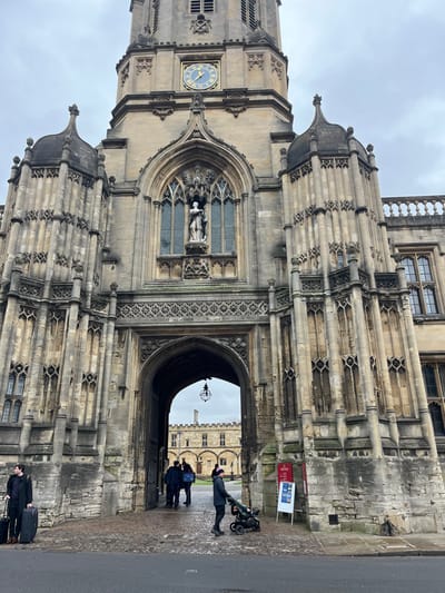 The image displays the Tom Tower and Tom Gate of Christ Church, Oxford, United Kingdom, under an overcast sky. The prominent stone structure features a central clock tower with a blue-faced clock displaying gold Roman numerals and hands, indicating approximately 10:15. Below the clock, a large arched window with intricate stone tracery contains a white statue of a robed figure. A grand, multi-layered arched gateway, Tom Gate, forms the primary passage through the building at ground level. The facade is adorned with intricate stone carvings, buttresses, and pinnacles. Through the archway, a historic stone quadrangle with additional institutional buildings and bare trees is visible, illuminated by a dark, ornate hanging lantern.
In the left foreground, a male subject, dressed in a dark coat and trousers, stands with two dark wheeled suitcases, looking towards the left side of the frame. In the right foreground, a female subject, wearing a dark jacket and a dark beanie with a pink pom-pom, pushes a dark stroller containing an infant or child, positioned next to a white A-frame information board labeled "Christ Church Oxford." Several additional figures are discernible walking within the quadrangle through the archway. The ground immediately in front of the archway consists of light-colored paved stone, bordered by a darker asphalt road in the extreme foreground.