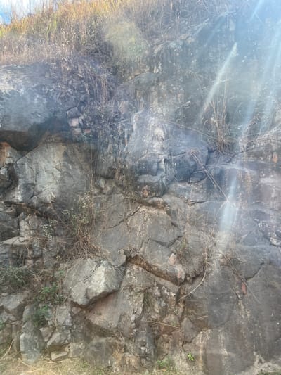 The image captures a rugged, weathered rock face, likely a section of a cliff or hillside, in a natural outdoor setting. The dominant features are varying shades of grey and dark-toned rock, characterized by numerous cracks, fissures, and distinct layered formations. Some parts of the rock appear to be damp or reflective, catching the light.
Sparse vegetation, primarily consisting of dry, light-colored grasses, thin branches, and small patches of green foliage, grows in crevices and along the contours of the rock face.
The scene is brightly lit by direct sunlight, which creates a prominent lens flare originating from the upper left portion of the frame, indicating a clear, sunny day. The light accentuates the textures and contours of the rock. No people, man-made objects, or visible text are present in the image. The setting suggests a natural landscape in a rural or semi-rural area, consistent with Thôn 8, Vietnam.