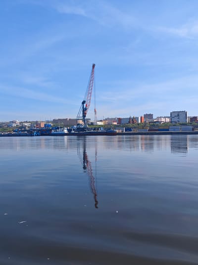 This image depicts a waterfront scene in Dudinka, Russia. The primary subjects are a large construction crane and a dock filled with shipping containers. Several boats are moored along the dock, including a white and blue vessel. In the background, a city skyline is visible, characterized by multi-story residential buildings and a church with a prominent dome.

The scene appears to be a busy industrial port, likely involved in shipping and cargo handling. The crane suggests ongoing construction or loading/unloading operations. The water in the foreground is calm, reflecting the sky, buildings, and the crane, indicating a peaceful, perhaps early morning or late afternoon setting.

The sky is a clear, bright blue with a few wispy clouds, suggesting fair weather. The sunlight casts distinct reflections on the water, illuminating the scene. The presence of numerous shipping containers points to a location involved in international trade or resource transportation, common for port cities like Dudinka.

There is no visible text in the image. The overall mood is one of industrial activity set against a backdrop of a functional, likely cold-climate, urban environment. The reflection of the crane and the cityscape creates a symmetrical and visually striking element in the composition.
