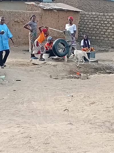 This image depicts an outdoor scene in Nata-Ala, Nigeria, centered around a manual water pump situated on uneven, sandy terrain. Six individuals and one light-colored dog are present.
From left to right: A man in a light blue caftan and dark trousers walks toward the right, appearing to hold a small object in his left hand. To his right, a woman stands with her hands on her hips, wearing a patterned short-sleeved top and a long patterned skirt. Adjacent to her, a woman in an orange top, patterned skirt, and red head covering operates the manual water pump, actively pulling its handle upwards. A large black tire rests against the right side of the pump structure, which is set on a low concrete base.
To the right of the actively pumping woman, another woman stands, dressed in a white short-sleeved top, a patterned skirt, and a red head covering. Further right and slightly behind the pump, a fifth woman, wearing a white top and a dark head covering, sits on the ground beside a blue plastic container. Colorful red and yellow patterned fabric is spread in front of her.
A light-colored, short-haired dog walks from right to left in the mid-ground, passing the water pump area. A white bucket is visible near the base of the pump. The background features structures with walls constructed from mud bricks or rough stone, topped by corrugated metal roofs. The ground throughout the scene is dry, light brown, and dusty, with some scattered debris. The scene is illuminated by natural daylight.