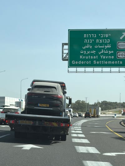 A forensic description of the scene reveals a bright daylight highway environment within the Hevel Yavne Regional Council, Israel. The primary subject is a dark-colored flatbed tow truck traveling on an asphalt highway with white lane markings. The tow truck carries a black Mercedes-Benz EQS 350 SUV, loaded rear-first onto its flatbed. The SUV displays an Israeli license plate "778-77-003" and has "EQS 350" visible on its tailgate. The tow truck itself bears an Israeli license plate "698-82-703" on its rear bumper.
The highway features multiple lanes, with the viewer's lane marked by a prominent white forward-pointing arrow. To the right, white chevron markings delineate a diverging or merging lane. Overhead, a large green highway sign mounted on a metal gantry structure provides directional information in Hebrew, Arabic, and English. The sign indicates directions to "Gederot Settlements" (associated with Road 4000, partially visible) and "Kvutsat Yavne" (associated with Road 4102), with an arrow implying a rightward turn or exit for these destinations. Kvutsat Yavne is a kibbutz located within the Hevel Yavne Regional Council.
Other vehicles are present on the highway, including a white Peugeot SUV partially visible behind the tow truck and a line of vehicles in the distance. Further down the road, a white truck is observed towing a yellow modular unit. The background includes sparse vegetation, utility poles, and industrial-like structures under a clear, light blue sky. No individuals are explicitly visible.