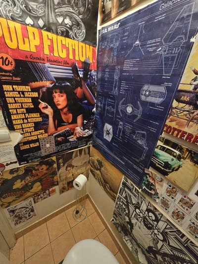 The image shows a collage of posters and other ephemera covering a wall in a room, likely a bathroom, given the presence of a toilet paper holder and toilet in the bottom right corner. The posters include a "Pulp Fiction" movie poster, "Star Wars" blueprints featuring the Millenium Falcon and TIE fighter, and various other images including illustrations, playing cards and comic book art. The posters are taped haphazardly to the wall, creating a dense, layered composition. The environment appears to be a small, cramped space, possibly a bathroom based on the presence of the toilet roll holder and toilet seat. The tiled floor and the perspective of the shot suggest it is a low-angle view taken from within the space. The location in Aubervilliers, France, is not evident in the image.