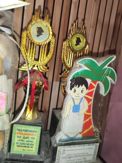 The image displays a cluttered indoor scene featuring multiple awards, predominantly trophies, against a wooden paneled background. Two main trophies are prominently visible, both heavily covered in dust and cobwebs.
The leftmost trophy features a golden design with a central circular emblem depicting a black silhouette of a person wearing a graduation cap. Below the golden top, red ribbons are tied around the trophy stem. A green label on its black base reads: "PIALA PENGHARGAAN" (Award Trophy), "WISUDA GABUNGAN XI" (11th Combined Graduation), "TKA - PLUS / TPA" (Kindergarten - Plus / Qur'an Learning Park), "LPP TKA - BKPRMI", "KEC. CILEUNGSI, KAB. BOGOR" (Cileungsi District, Bogor Regency), "TAHUN PELAJARAN 2009 - 2010" (Academic Year 2009 - 2010), and "TGL. 21 JUNI 2010" (Date: 21 June 2010).
The rightmost trophy has a similar golden design at its top, also featuring a graduation cap silhouette. Its base is partially obscured by a plastic-wrapped, cut-out figure of a young boy with dark hair, wearing a white shirt and blue overalls, standing next to a cartoon palm tree. A white label on this trophy's base reads: "HIMPAUDI KECAMATAN CILEUNGSI" (HIMPAUDI Cileungsi District), "PERAGAAN MANASIK HAJI" (Hajj Ritual Simulation), "PAUD SEKECAMATAN CILEUNGSI" (Early Childhood Education across Cileungsi District), and "Bundok Gede, 29 Oktober 2019".
A third, partially visible object on the far left appears to be another award or decorative item, also covered in dust and plastic, with a glimpse of a pink artificial flower. The overall environment suggests long-term storage or neglect, characterized by significant dust accumulation and cobwebs on all displayed items. The location of the events referenced on the trophies is specified as Cileungsi District, Bogor Regency, Indonesia.