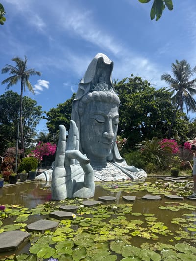 The image shows a large, grey statue of a deity, possibly a Buddhist figure like Guanyin, emerging from a pond filled with lily pads and lotus flowers. The statue's head is prominent, with closed eyes and a serene expression. One of its hands is raised in a gesture, with fingers extended. The statue is surrounded by lush greenery, including palm trees and flowering bushes in vibrant pinks and purples. The pond has stepping stones leading across it. In the background, a person wearing a white top and light-colored shorts is visible, standing near the edge of the pond. The sky is blue with scattered white clouds, and the overall setting appears to be a temple or garden. The statue is located at the Long Son Pagoda in the city of Hòa Đa, Vietnam.