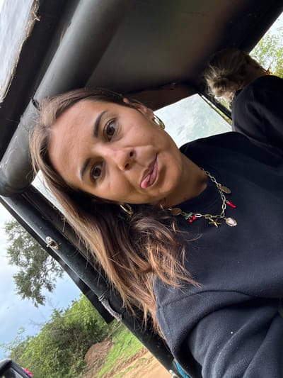 A woman is seen from a close, upside-down perspective inside an open-top vehicle. She sticks her tongue out playfully. She has brown hair, brown eyes, and gold hoop earrings. She wears a black sweatshirt and a long, ornate charm necklace. Outside the vehicle, green vegetation and a partly cloudy sky are visible. Another person with blonde hair tied up in a bun is seen looking into the frame. The photo appears to be taken on a safari tour.