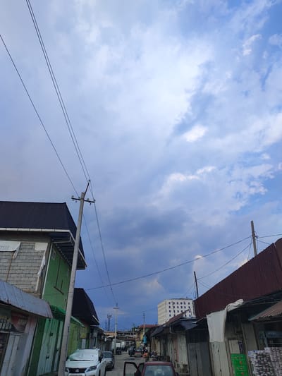 The image depicts a street scene in Tursunzoda, Tajikistan, under a cloudy sky. The perspective is from street level, looking upwards and down a street lined with buildings and shops. On the left, a green building with a dark roof is partially visible, adjacent to a building with grey brickwork. A utility pole with multiple wires extends upwards into the sky. Further down the street, several shops with corrugated metal roofs are visible. A white SUV is parked on the right side of the street, and other vehicles are present further in the distance. A modern, multi-story white building with numerous windows stands prominently in the background, contrasting with the older structures. The sky is a mix of white and dark grey clouds, suggesting overcast or possibly rainy weather.