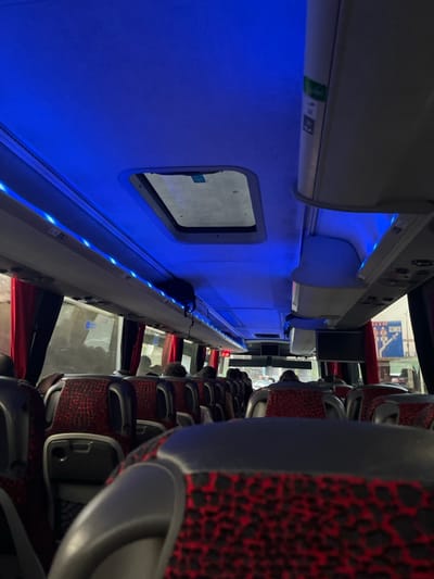 The image shows the interior of a bus, likely a tour bus, from the perspective of a passenger seated near the back. The bus has blue LED lights and an overhead hatch. Red curtains are visible outside the windows. There are passengers seated in rows throughout the bus. The bus is stopped, as a sign is visible through the window behind the driver's seat. It is likely located in the city of Joinville-le-Pont, France, based on the coordinates.