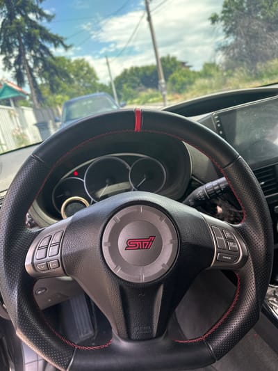 The image shows the interior of a car, focusing on the steering wheel with an "STI" logo. The steering wheel is black with red stitching. The car's dashboard and a glimpse of the outside environment are visible through the windshield. The environment shows trees, a sky with light cloud cover. The location is indicated as Golyanovtsi, Bulgaria.