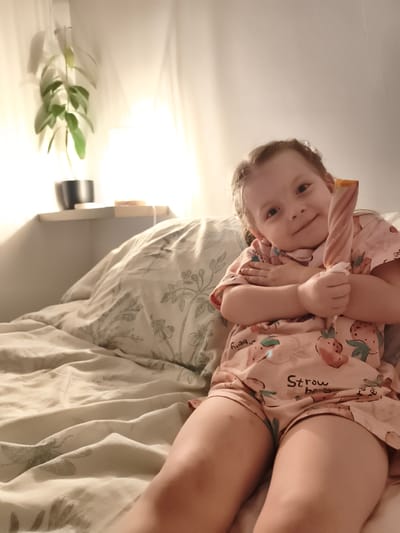 A young girl is sitting on a bed, holding a colorful popsicle. She is wearing a pink pajama set adorned with strawberries and the text "Strawberry". The bed is covered with a light-colored duvet featuring a floral and dragonfly pattern. In the background, a shelf holds a potted plant and a small wooden object. A soft light emanates from a lamp, casting shadows on the wall, suggesting it might be evening or a dimly lit room. The girl has a gentle smile and appears content. The setting seems to be a bedroom, possibly in Lagny-sur-Marne, France.