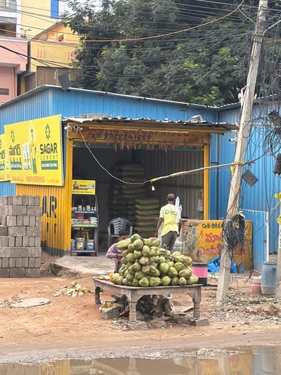 The image depicts a street scene in Shamshabad, India, likely near a construction supply store. In the foreground, a pile of green coconuts is displayed on a wooden table. Behind the coconuts, a person is visible, possibly a vendor. To the left of the coconuts, stacks of grey cinder blocks are piled up. The central part of the image features a blue corrugated metal building with a yellow awning and signage that reads "SAGAR CEMENT." The store appears to be selling construction materials, with stacks of cement bags visible inside. A man in a yellow t-shirt is walking away from the store. Electrical wires are prominently strung across a utility pole on the right side of the frame. The ground is muddy and appears to have puddles, suggesting recent rainfall. Buildings and trees form the background.