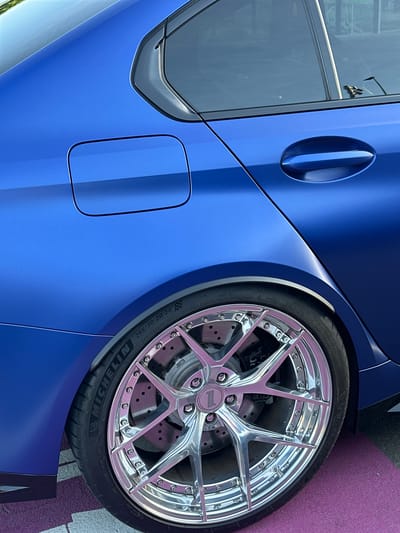 A close-up shot of the rear driver's side of a blue car is depicted. Key details include the car's door handle, fuel door, and a prominent chrome wheel and tire combination. The tire is branded as MICHELIN, with markings indicating "265/30ZR20 (99Y) EXTRA LOAD" and "285/30ZR20". The presence of road markings, specifically purple paint, is visible beneath the car. There are no people visible in the frame. The location, Volketswil, Switzerland, is mentioned but not visibly confirmed in the image.