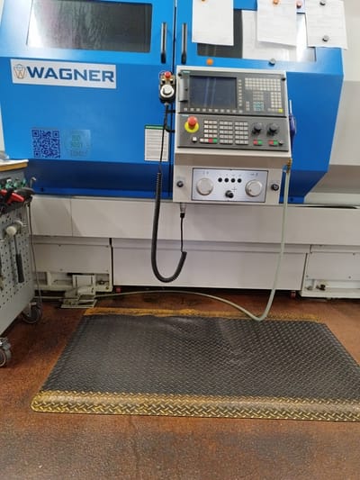 This image shows the interior of a workshop or industrial setting, likely in Bremerhaven, Germany, with a prominent blue industrial machine as the central subject. The machine features a control panel with a screen, various buttons, and knobs, as well as a "WAGNER" logo. Attached to the machine is a coiled black cable and a flexible light-colored tube, suggesting it is a piece of automated equipment. To the left of the machine, a mobile tool cart filled with various tools, including hammers and pliers, is partially visible. In front of the machine, a black anti-fatigue mat with a yellow-bordered edge is placed on the floor. The floor itself appears to be made of concrete or a similar industrial flooring material, with some reddish-brown staining. The overall environment appears functional and utilitarian, consistent with a manufacturing or repair facility. There are no people visible in the image. The context of Bremerhaven is not explicitly stated but is provided as information about the image's location.