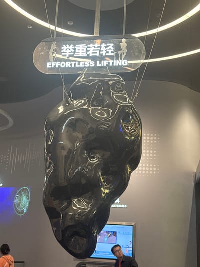 An irregularly shaped, large, dark, highly reflective sculpture is suspended from the ceiling in an indoor exhibition space in Guangming District, China. The sculpture's surface exhibits distorted reflections of the surrounding environment, including overhead lighting. Above the sculpture, a rectangular sign with rounded corners displays the Chinese characters "举重若轻" and the English phrase "EFFORTLESS LIFTING" in white text. The sign shows visible smudges or residue. The ceiling is dark, featuring integrated linear light strips. The background walls are light gray, displaying digital projections or screens; one on the left shows abstract patterns and a circular diagram, while another to the right features colorful imagery. The word "MATERIALS" is partially visible on the wall. In the lower right of the frame, a male individual wearing glasses and a dark shirt looks upward and to the left. In the lower left, the back of a person with light brown hair, wearing an orange top, is partially visible. The scene depicts a modern, well-lit public or exhibition area.