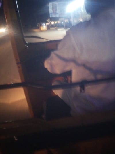 The image depicts the interior of an auto-rickshaw at night, as seen from the passenger's perspective. A person wearing a white shirt is visible in the driver's seat, their back and shoulder dominating the right side of the frame. The scene is dimly lit, with streetlights and vehicle headlights casting glows on the road and the auto-rickshaw's interior.

Outside the vehicle, a street scene is visible through the windshield and side windows. Headlights of other vehicles can be seen on the left, indicating movement on a road. A yellow auto-rickshaw or similar vehicle is parked or moving in the distance, along with other structures that appear to be shops or buildings lining the street. A bright, possibly artificial light source is present in the background, illuminating a large advertisement banner or sign. The image on this sign is blurred but appears to feature a person's face and some text.

The overall impression is that of a typical night drive through a populated area. The time of day is clearly night due to the darkness and artificial lighting. The weather appears clear as there are no visible signs of rain or fog. The location cues suggest an urban or semi-urban environment in Nigeria, specifically Baccure Kofare, as indicated by the metadata. No specific emotions are conveyed by the subjects in the image due to the blurriness and focus on the environment and the driver's silhouette. The primary activity is transportation, with the auto-rickshaw likely in motion or stopped at a light.