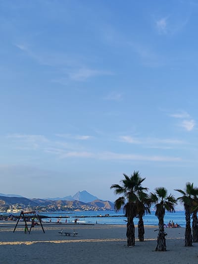 This media file depicts a sandy beach with palm trees in the foreground, overlooking a calm blue sea. In the background, a coastal town with buildings and boats lines the shore, with a prominent mountain rising behind it. Several people are visible on the beach and in the water, enjoying a leisurely day. Some are sunbathing, others are swimming, and children appear to be playing on a swing set and a seesaw. The sky is a clear blue with a few wispy clouds, suggesting it is daytime, likely late afternoon given the warm lighting. The overall atmosphere is peaceful and relaxed, conveying a sense of a typical summer day at a seaside resort. There is no visible text in the image.