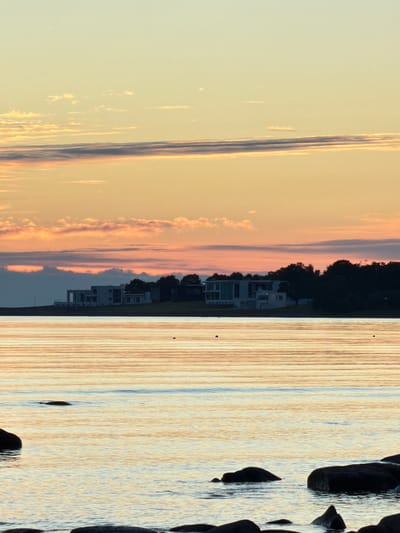 A vertical, full-frame shot captures a tranquil sunset over the Baltic Sea at Haabneeme alevik, Estonia. The sky displays a gradient of soft yellow and peach hues, with delicate, elongated clouds stretched across the horizon. Below the sky, a thin, dark band of clouds sits just above a row of modern, white-and-glass houses situated on a gentle slope. These residences are bordered by dark, silhouetted trees. The sea in the foreground reflects the warm colors of the sky, creating horizontal bands of light and shadow on its surface. Several dark, wet rocks are partially submerged in the water along the bottom edge of the frame, with more rocks visible further out in the water. Two small, dark buoys or markers are visible in the distance on the water. The scene is devoid of any people.
