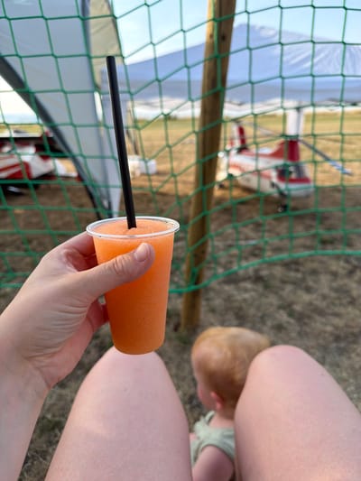 In Becherbach, Germany, a person's hand holds a clear plastic cup filled with an orange, frozen beverage with a black straw. The cup is positioned in the foreground, in front of a green mesh fence. Behind the fence, two children are visible. One child, with blond hair, is seated and appears to be looking away from the camera. The other child, in the background, is out of focus. The scene suggests an outdoor setting, possibly a recreational area or event, with a tent and what appears to be a model airplane visible in the distance. The overall lighting indicates daytime.