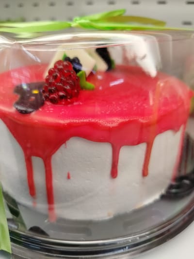 A close-up, slightly out-of-focus shot shows a white cake in a clear plastic container. The cake is covered with a bright red, glossy glaze that drips down the sides. On top of the cake, there are decorative elements including a red, berry-shaped piece that appears to be made of candy or gelatin, and what looks like a black berry. A bright green ribbon is visible in the background, tied in a bow, suggesting a gift or celebration. The cake is likely on display, possibly in a bakery or store. The image does not provide any specific context or details related to Yashkino, Russia.