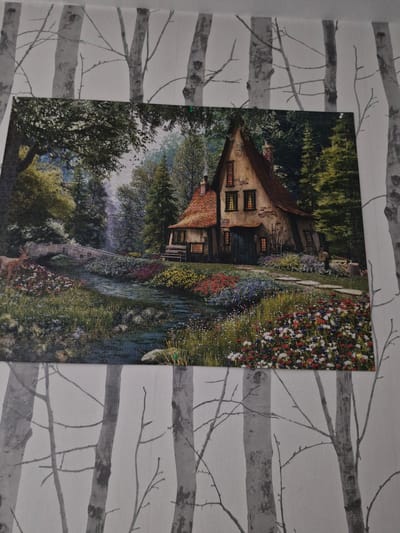 The image shows a puzzle depicting a whimsical cottage in a lush, wooded landscape. A small stone bridge crosses a gentle stream, which flows through a meadow filled with colorful wildflowers. A deer stands near the stream, and the cottage itself has a red-tiled roof and glowing windows, suggesting a cozy interior. The puzzle is placed against a backdrop of wallpaper featuring a pattern of bare birch trees. There is no indication of the city of Katowice, Poland, in the image.