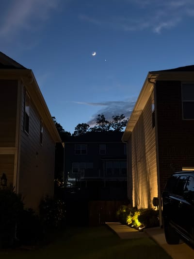 The image depicts a twilight scene in a suburban neighborhood, possibly in Buford, Georgia, United States. The sky is a deep blue transitioning to lighter shades with some thin clouds. A crescent moon and a bright star, likely Venus, are visible in the upper portion of the sky. The foreground is dominated by the sides of two houses, one on the left with beige siding and the other on the right with red brick. Both houses have visible windows and architectural details. Between the houses, a dark silhouette of another house is visible in the background, along with trees. The right side of the image shows a dark SUV parked in a driveway and landscaping illuminated by yellow uplighting. The overall impression is a quiet residential evening scene.