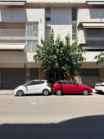 This is an eye-level, slightly tilted shot of a residential building facade in Vilanova i la Geltrú, Spain. Two cars, a red hatchback and a white hatchback, are parked vertically on the street beside the building. The red car is closer to the building, and the white car is in front of it.
The building is a light beige color with multiple floors and windows protected by white metal bars. Some windows have white blinds or awnings. A large tree with lush green leaves is growing in front of the building, obscuring some of the lower floors. The street is paved with asphalt and is in shadow from the building and possibly another structure not fully visible.
The sun is casting strong shadows, suggesting it is either late morning or early afternoon. The light is bright and the shadows are distinct, indicating clear weather. The overall scene is calm and urban, with no people visible. There is some graffiti on the grey metal shutter of the ground floor entrance of the building, but it is not legible.
