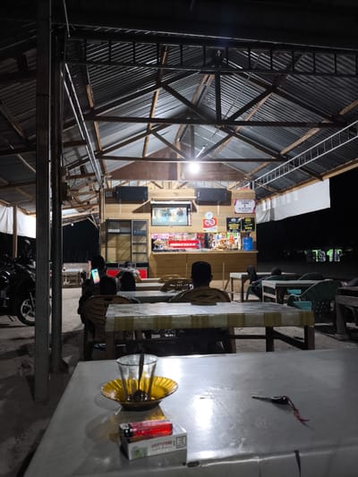 The image depicts an outdoor eatery at night in Ujongreuba, Indonesia. The structure has a corrugated metal roof supported by metal poles and wooden beams. Several tables and chairs are arranged outdoors. In the foreground, a table is covered with a plastic tablecloth and holds a glass of tea, a yellow saucer, a cigarette lighter, and a pack of Lucky Strike cigarettes. In the background, a counter is visible with shelves stocked with various items, including drinks and snacks. A television screen is mounted above the counter. Several people are seated at tables, some facing away from the camera. A motorcycle is parked on the left side of the image. The overall scene suggests a casual dining or gathering place.