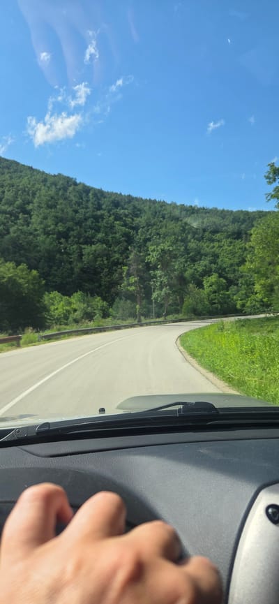 The image, taken from inside a vehicle, captures a scenic drive along a winding road through a lush, forested landscape. The perspective is from the front passenger side, looking out the windshield towards the road ahead and the surrounding nature.

On the right side of the frame, a person's hand is visible, resting on what appears to be the car's dashboard or door panel. Parts of the car's interior, including the windshield wiper and a dark grey dashboard, are prominent in the foreground.

The asphalt road curves gently to the right, bordered by green grassy verges. A white solid line is painted on the road, indicating a lane or edge. Beyond the road, dense green forests cover rolling hills or mountains, stretching across the middle ground and background of the image. The trees appear deciduous, showcasing vibrant green foliage.

Above the treeline, a clear blue sky dominates the upper portion of the scene, dotted with a few scattered, wispy white clouds. The bright illumination suggests it is daytime, likely midday or afternoon, with good weather conditions.

The overall scene depicts a tranquil journey through a natural environment, potentially in the vicinity of Debnevo, Bulgaria, as indicated by the context. No specific text is visible in the image.