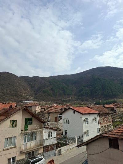 This image depicts a residential area in Krupnik, Bulgaria, with a mountainous backdrop under a partly cloudy sky. The scene is viewed from an elevated perspective, looking down onto a cluster of buildings and a street.

In the foreground and mid-ground, multiple multi-story residential buildings are visible, characterized by predominantly terracotta-tiled roofs. Building exteriors vary in color, including beige, white, and light brown. One prominent beige building on the left features multiple white-framed windows and a distinct green main door, along with a balcony with metal railings. Directly opposite, a newly constructed or renovated white building stands out with clean white walls, multiple white-framed windows, and two visible exterior air conditioning units on its side. Other buildings in the immediate vicinity show signs of age, with some featuring exposed brickwork.

A paved street runs through the scene, with a silver sport utility vehicle (SUV) partially visible parked on the street in the lower-left quadrant. Power lines and utility cables are strung between poles and across buildings.

The background is dominated by a large, steep mountain range. The mountains display a mix of vegetation, with sections of bare trees indicating either a dormant season (late winter/early spring) or deciduous forest, interspersed with darker green evergreen sections. The sky above is light blue with scattered white, wispy clouds. No individuals or explicit actions are discernible within the frame.