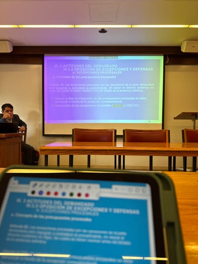 This image depicts an indoor scene within a classroom or conference room identified as "Salon 211" in Tizapan, Mexico. The perspective is from a seated individual, viewing a presentation.
In the foreground, a dark green-cased tablet-like device is partially visible, its screen displaying a digital document with a blue background and white text. The document's header, "III. 3 ACTITUDES DEL DEMANDADO III.3.5 OPOSICION DE EXCEPCIONES Y DEFENSAS A. EXCEPCIONES PROCESALES," mirrors the content on the main projector screen. An annotation toolbar with colored circles (black, purple, red, green) is present at the top of the tablet's display.
In the background, a large projector screen dominates the center, displaying a presentation slide in Spanish with a blue-to-green gradient background. The slide's title is identical to the tablet's header. Below the title, the text "a. Concepto de los presupuestos procesales" and legal definitions are visible, including "Artículo 62. Las excepciones procesales son las oposiciones de la parte demandada..." The top center of the projected image clearly shows "Nombre Sala : Salon211".
To the left of the projector screen, a male presenter is seated behind a dark wooden lectern. He is wearing a dark suit jacket over a white shirt, with a dark laptop open in front of him. His right arm is raised, and his index finger is extended, suggesting he is actively gesturing or pointing while speaking.
In front of the projector screen, a long, polished wooden table with dark metal legs extends across the frame. Several chairs with dark red upholstered backs and seats are arranged behind this table. The room's ceiling features linear fluorescent lighting fixtures and a white access panel. A white air conditioning unit is mounted on the right wall. The overall lighting is bright and even.