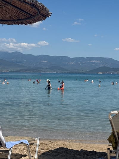 In the city of Lichada, Greece, a scene unfolds at a beach on a sunny day. The turquoise water stretches towards a distant mountain range under a clear blue sky dotted with a few wispy clouds. In the foreground, a straw umbrella shades a section of the beach, and a beach chair with a blue towel draped over it is partially visible. The water is populated with numerous people swimming and enjoying the sea. Several individuals are wading in the shallows, while others are further out, some with swim rings. The overall atmosphere is one of leisurely summer activity at the seaside.