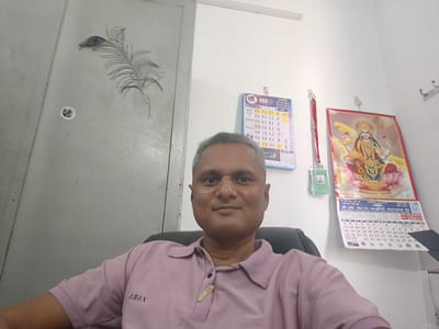 A medium shot captures a man with short, gray hair, seated in a black office chair and facing the camera. He is wearing a light pink polo shirt. Behind him, a gray metal door with a peacock feather decoration is visible on the left side. On the wall behind him, there is a calendar hanging, along with a poster of a deity. There is no indication of the location being Dhandhuka, India in the provided context.