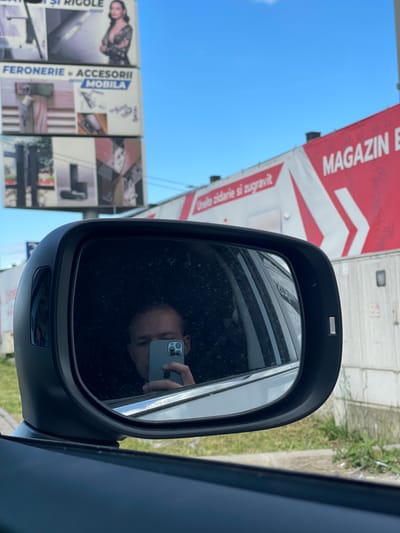 The photograph appears to be taken from the interior of a vehicle, focusing on the side mirror. The reflection in the mirror shows a person holding a smartphone, ostensibly capturing the image itself. The reflection also reveals elements outside the vehicle, including commercial signage and parts of a building. The signage includes the words "MAGAZIN" and "FERONERIE," with additional text in Romanian. Specific signs for "Feronaerie si Accesorii Mobila" (Hardware and Furniture Accessories) can be seen. Given the Romanian text, the location is most likely in Romania. Based on the description and information available, the photograph shows commercial establishments in the city of Afumați, Romania.
