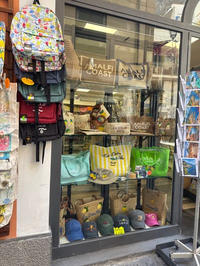 The image shows a storefront in Pogerola, Italy, displaying a variety of merchandise. The window is filled with colorful backpacks, woven tote bags, hats, and postcards. Several items are branded with "Amalfi Coast" and "La Dolce Vita," suggesting the location is a tourist-oriented shop selling souvenirs. The backpacks are stacked vertically, featuring a playful print with scooters and beach scenes. Woven straw bags, some with embroidered lettering and designs of lemons, are arranged on shelves. A selection of baseball caps with various slogans and graphics is displayed on a lower shelf. Postcards depicting scenic views of coastal towns are mounted on a rack to the right. The reflection in the window shows buildings and a balcony, typical of a Mediterranean town. The street in front of the store appears to be paved, with a granite-like texture on the lower part of the building.