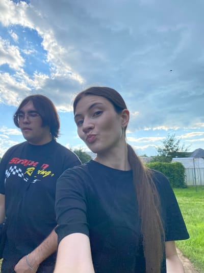 Two people are posing for a selfie outdoors. The person in the foreground on the right has long brown hair and is wearing a black t-shirt, making a kissing face at the camera. Behind them and slightly to the left, another person with brown hair and glasses is visible, also wearing a black t-shirt with yellow and red text that appears to say "Strange Racing". They are both looking towards the camera. The background consists of a blue sky with scattered white clouds and greenery, suggesting a daytime setting. A small insect is visible in the sky above the two individuals. The scene appears to be a casual outdoor gathering in Novyi Ural, Russia, possibly during the daytime with fair weather.