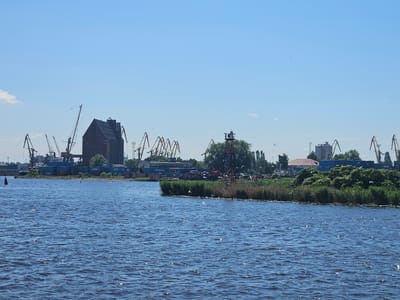 The image shows a wide, blue body of water with moderate waves reflecting sunlight. Along the far bank, there is a busy port scene under a clear blue sky. Several large cranes, some yellow and some red, dominate the skyline. A tall, dark brick building with a steep roof stands prominently in the middle ground, flanked by more industrial structures and shipping containers. Green trees and reeds line parts of the shoreline. Numerous white birds, likely seagulls, are scattered across the water and in the air. A single red buoy is visible in the water to the left of the frame. The overall impression is one of industrial activity and natural elements coexisting.
This scene is likely a view of the Pregolya River within the port of Kaliningrad, Russia. Kaliningrad is a major port city situated on the Baltic Sea, with the Pregolya River serving as a key waterway for its maritime activities. The presence of cargo ships, cranes, and industrial buildings is characteristic of this area.