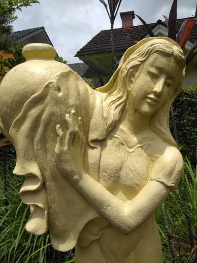 A light-yellow, granular-textured statue depicting a female figure carrying a large urn is positioned outdoors in Ba Nwet Kone, Myanmar. The statue's subject has a downward-tilted head, eyes cast low, and visible long hair. A large, round urn rests on her left shoulder, partially enveloped by draped fabric. Her right arm is bent, holding the urn, with the left hand also in a supportive posture. She is dressed in a short-sleeved garment. The background consists of an overcast sky above two partially visible buildings with dark, pitched roofs; one building on the right features a red brick chimney and green wall sections. Dense green foliage, including tall grasses and shrubs, occupies the foreground and mid-ground around the statue, with some reddish-leaved plants also present. The scene is under natural daylight.