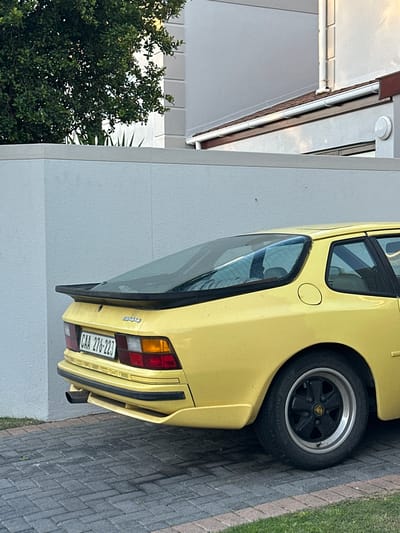 The image depicts the rear of a yellow Porsche 944 parked near a building in Blouberg, South Africa. The car has a license plate that reads "CAA 276-223". The environment includes a brick walkway, a building with white walls, a green tree.