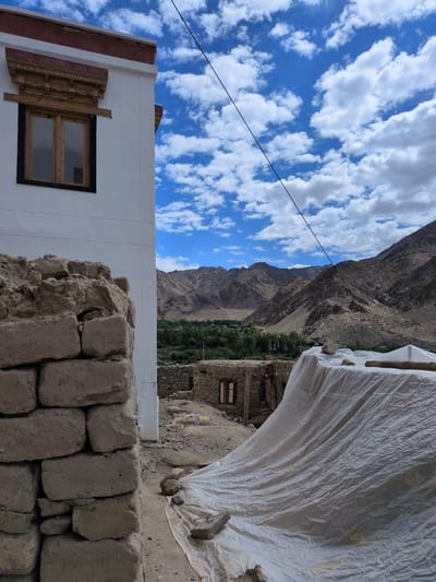 An exterior, eye-level, medium shot captures a section of a white building in the foreground, with a stone wall to its left. The building features a wooden-framed window adorned with intricate carved wooden detailing above it, and a red trim along the roofline. To the right of the building, a white tarp is draped over a structure, with a log and stones resting on top. In the mid-ground, there are partially constructed stone buildings with windows. Beyond these, a lush green valley with trees is visible, nestled between rugged, brown, mountainous terrain. The sky above is blue and filled with scattered white clouds. A thin black wire stretches diagonally across the sky from the top left to the middle right of the frame. The scene appears to be located in the city of Choma, India.