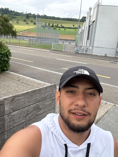 The image is a selfie of a man wearing a black "The North Face" cap and a white sports shirt. He has a beard. The background shows an asphalt road with yellow and white lane markings, a field, a building with visible pipes, and a fence. A transmission tower is visible on the horizon. The man is sitting on a wooden bench. The location is likely Lausen, Switzerland, given the landscape and the presence of the transmission tower, similar to those found in the area.