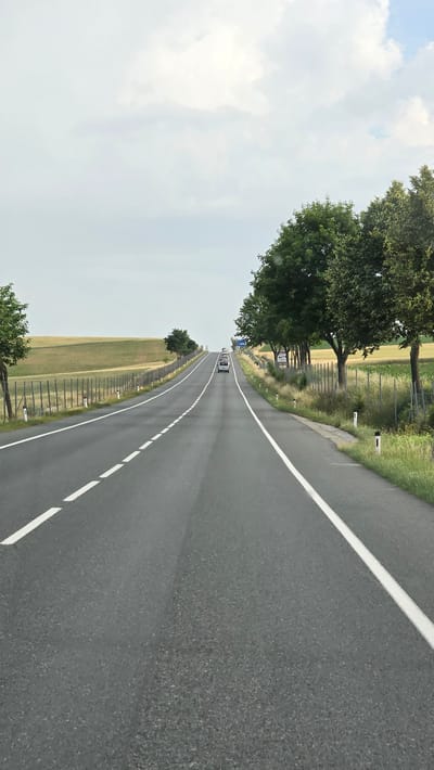 The image depicts a view from a vehicle traveling on a two-lane asphalt road that curves slightly to the left. White lane markings are visible on the road surface. To the left of the road, a grassy field with a wooden fence runs along the edge. Beyond the field, rolling hills can be seen. To the right of the road, a line of trees and vegetation is present, with a metal fence in between the road and the trees. Several white posts with black tops are intermittently placed along the edge of the road. In the distance, on the road, a line of vehicles, including what appears to be a white van and several other cars, are traveling in the same direction. The sky is overcast with white and gray clouds. The scene is consistent with a rural or semi-rural highway setting, likely in the vicinity of Poysdorf, Austria, given the landscape and road infrastructure.