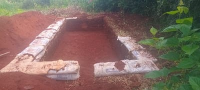 The image shows a rectangular pit dug in the ground in Rukenya, Kenya, located at the coordinates -0.559 latitude and 37.351 longitude. The pit is lined with several stacked bags of cement, creating a square perimeter. The ground surrounding the pit is red clay and there is a small green plant in the bottom right corner of the image. The pit appears to be in the process of being constructed. The bags are arranged in a straight line along the sides of the pit. The image was taken from a horizontal angle, looking down onto the pit.