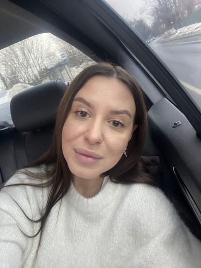 The image displays a female subject taking a selfie from within a vehicle, situated in what appears to be Vysokovo, Russia. The subject, a female with long, straight dark brown hair parted in the middle, light complexion, and brown eyes, is depicted from the upper chest up. She wears a white, chunky knit sweater, a thin silver chain necklace, and a small, silver-colored cross-shaped earring in her right ear. Her expression is neutral, with her lips slightly parted.

The interior of the vehicle shows a dark-colored seat headrest to her left and a dark-colored interior panel to her right. The background, visible through the vehicle's windows, portrays an outdoor scene during what appears to be winter or early spring. Water droplets are visible on the window glass. The environment consists of a wet asphalt road, flanked by piles of snow. Bare trees line the roadside. A blue and white bus stop sign, featuring a stylized bus icon, is partially visible through the rear window on the left side of the frame. Further along the road on the right, a green fence or wall is discernible, and distant vehicles are faintly visible. The sky is overcast and grey.