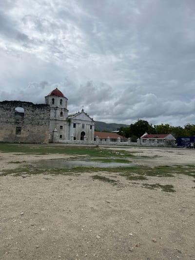 The image shows the historic Immaculate Conception Parish Church in Oslob, Philippines. The church, constructed with white stone and featuring a red dome, is situated next to a ruined stone structure on its left. In the foreground, a large open area of gravelly ground with patches of green grass and puddles of water suggests a recent rainfall. Behind the church, several smaller buildings with red roofs are visible, and further in the background, trees and rolling hills can be seen under a cloudy sky. A blue truck is parked to the far right of the frame.