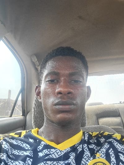 An image taken in the city of Nwofia, Nigeria, shows a single male individual seated in the front passenger compartment of a vehicle. The subject, an adult male with dark skin and short, dark hair, looks directly into the camera with a neutral expression. His face exhibits a slight sheen, possibly moisture. He wears a dark blue or black short-sleeved shirt with a yellow collar and yellow accents. The shirt is patterned with repeating white text, prominently displaying "INTER," and features a circular yellow emblem on the lower right side. A thin, gold-colored chain is visible around his neck.
The vehicle's interior features a light brown or beige headliner and similarly colored, somewhat worn upholstery on the seats, including a striped headrest behind the subject. The rear passenger seats are visible, appearing light-colored and showing signs of wear. Through the left-side window, a bright, hazy outdoor environment is discernible, along with a partial view of a rudimentary outdoor structure, possibly a fence or wall, made of rough materials. The overall lighting suggests daylight conditions. No other individuals or significant objects are present within the frame.
