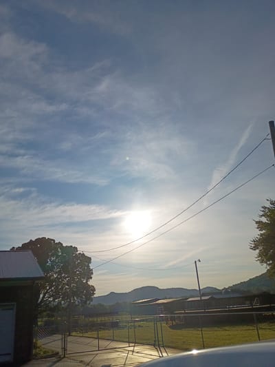 The image depicts a sunlit outdoor scene, likely during the late afternoon given the warm glow and long shadows. The sky is a prominent feature, filled with wispy white clouds that streak across a bright blue expanse. The sun, a brilliant white orb, is partially obscured by clouds, creating a lens flare effect and casting a warm light over the landscape.

In the foreground and mid-ground, elements of human infrastructure are visible. Two wires stretch diagonally across the frame, suggesting utility lines or perhaps part of a fence. A single-pole telephone or utility pole stands vertically, with some equipment attached near its top. To the right, a chain-link fence encloses an area with a grassy field, possibly a park or sports field. A small, white-roofed structure, likely a shed or utility building, is situated in the lower left portion of the image.

In the background, rolling hills form a natural backdrop, bathed in the soft light of the setting sun. The overall impression is one of tranquility and open space, with a hint of rural or suburban atmosphere. No people are visible in the image. The weather appears clear with scattered clouds, and the time of day is consistent with a late afternoon or early evening. The location context of Berea, USA, suggests this scene could be in a park, a residential area, or near a recreational facility. There is no visible text in the image.