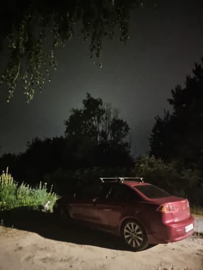 An exterior, low-angle, medium shot captures a red sedan parked on a dirt path at night in Szczaki, Poland. The vehicle, facing left, has a roof rack and a license plate reading "WPP 5Z04". Overgrown green vegetation and trees form a dark backdrop against a hazy, illuminated sky. The foreground shows a rough, sandy surface with scattered debris. The lighting suggests an outdoor, possibly rural, setting with ambient light from an unseen source.