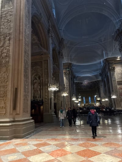 The image depicts the interior of a large, ornate church, identified as the Cattedrale di San Giorgio Martire (Ferrara Cathedral) in Ferrara, Italy. The perspective is from a slightly low angle, looking down a nave or side aisle towards the altar area in the distance.
**Environment:**
The cathedral features towering, massive columns or pilasters with elaborate relief carvings and decorative elements, including niches with statues. At least one light-colored statue of a draped figure is visible in a niche on a column to the left. The high, vaulted ceiling is partially obscured by a light-colored safety netting, stretching horizontally across the upper sections, indicating ongoing restoration or structural protection. Multiple ornate, multi-tiered chandeliers with clear glass or crystal components and numerous light bulbs hang from the ceiling, providing illumination. The floor in the foreground is a checkerboard pattern of alternating light (cream/white) and reddish-brown square tiles, appearing somewhat reflective. In the background, beyond more chandeliers and architectural features, stained-glass windows in shades of blue and yellow are visible at the far end of the nave, where rows of dark pews or chairs are arranged, suggesting an active worship space.
**People:**
Five individuals are clearly visible in the mid-ground. One person, wearing a dark blue puffy coat, black trousers, and a bright pink scarf or hat, walks towards the viewer, slightly right of center. Three other individuals are walking away from the viewer, further into the cathedral: one in a light beige coat, light blue jeans, and a green scarf; and two others in dark coats. An additional indistinct figure is visible further in the background. All individuals are dressed in winter-appropriate outer clothing. No direct interactions between the visible individuals are discernible; they appear to be moving through the space independently.