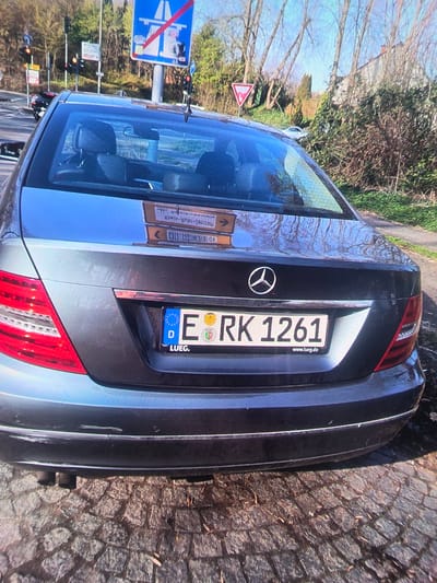 A dark gray Mercedes-Benz sedan is parked on a cobblestone street in front of a building and a wooded area. The car's rear is visible, including its taillights, license plate, and exhaust pipes. The license plate reads "RK 1261" and has a "D" emblem, indicating it is registered in Germany. A small sticker with the text "www.lueg.de" is also present on the license plate. In the background, traffic lights and road signs are visible, suggesting a road intersection. The environment appears to be outdoors, under daylight.